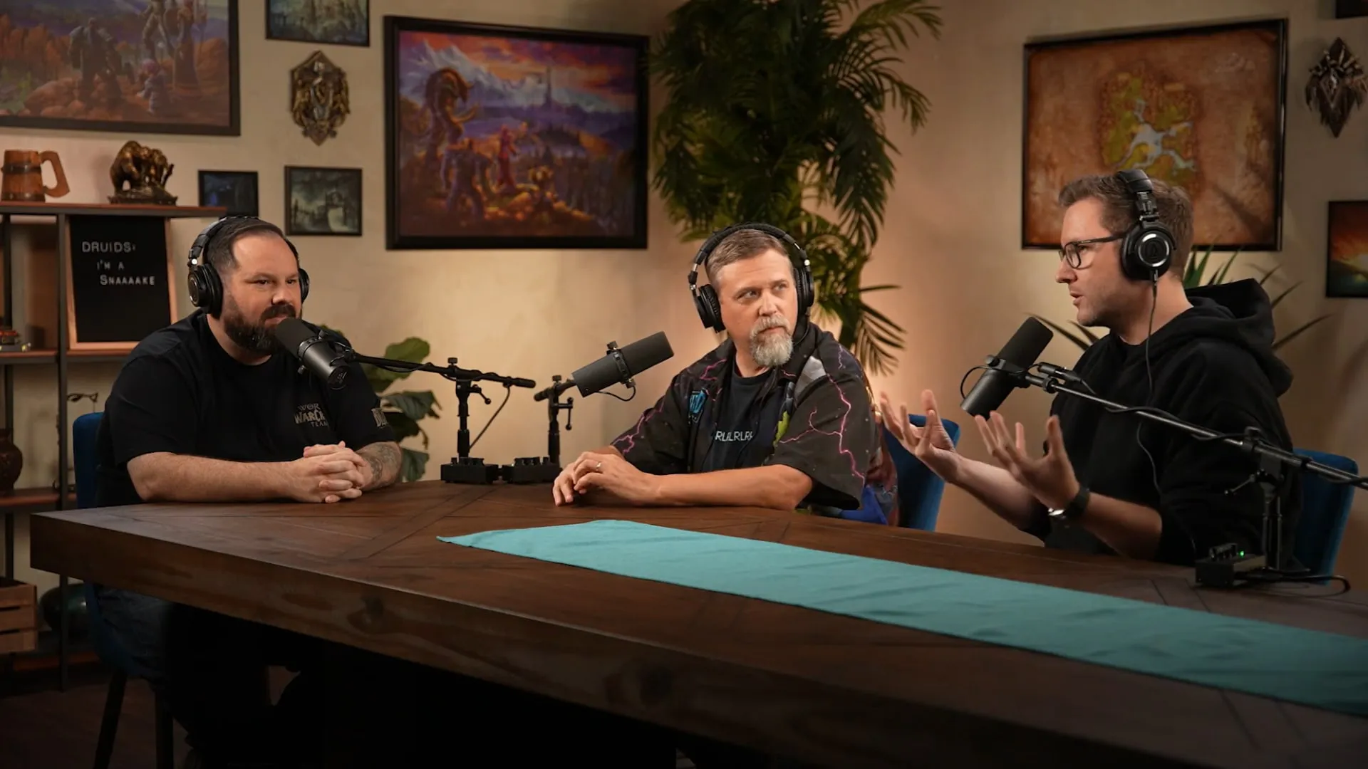 Three Blizzard developers with headphones at a wooden table with microphones, in a studio with World of Warcraft paintings and a map on the wall